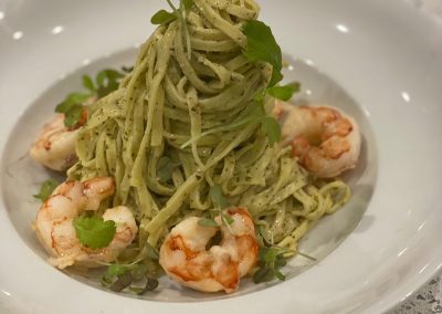 Shrimp pesto dish with pasta covered in green pesto, spun into a tall mound on a white plate. Shrimp on the perimeter of the shallow bowl.