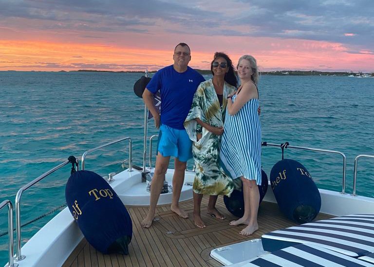 Three adults standing on the bow of Top Shelf on their yacht charter. Background: blue water, cloudy sky with pink and orange sunset.